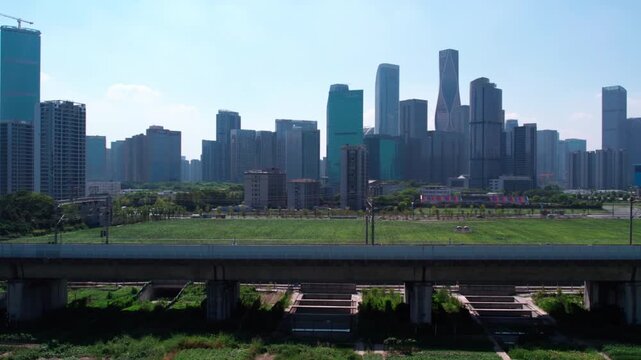 A modern urban view featuring a skyscraper skyline, an overpass, and a large green open space in the foreground under clear daytime sky.