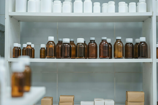 Rows of amber glass medicine bottles and white plastic containers on shelves in a pharmacy storage room with cardboard boxes on the bottom shelf