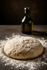 Fototapeta premium Raw dough rests on dark table with flour dusting. Green glass bottle of oil stands in background. Preparing fresh baked goods for home baking.