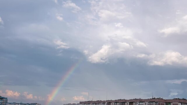 A vibrant rainbow forms in the cloudy sky as storm clouds move rapidly over urban rooftops in time-lapse. Concept of hope, good omens, optimism, divine signs, and natural beauty.