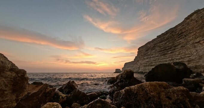 Golden sunset rays illuminate limestone rock formations along the turbulent Black Sea coastline during twilight. Low angle wide shot.