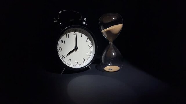 Vintage alarm howing 8 o'clock and hourglass with falling sand on black background. Concept of morning, work time, new day and time management.