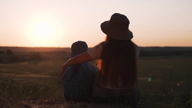 Mother holds child while sitting at sunset in countryside. Woman embraces boy during golden hour outdoors. Family bonds at sunset together. Mother and child embrace in nature. Boy with mother at dusk.
