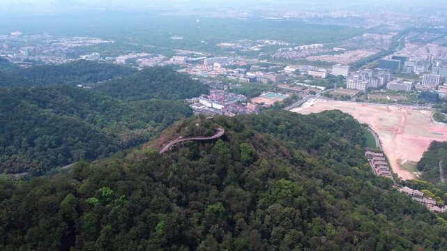This is an aerial landscape showing a forested hill with a winding path, overlooking a distant urban cityscape and natural surroundings.