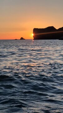 Golden sunset glowing behind rocky cliffs over calm ocean waters. Fixed perspective shot capturing evening tide gently rolling onto sandy beach shore.