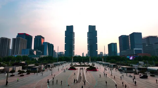 A spacious urban plaza with walking people, surrounded by modern skyscrapers under a pale sky. Tree belts line the edge of the open public space.