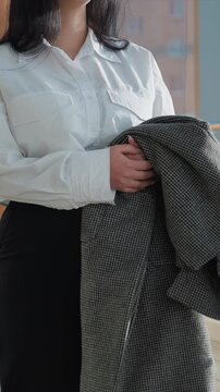 Young woman walks indoors holding gray houndstooth coat wearing. Woman turns her head slightly while standing near large windows. Wooden pillar is visible in background