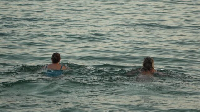 Sea bath by two women swimming in open water during evening twilight. Static wide shot.