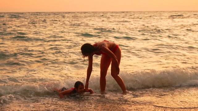 Stunning coastline sunset captures mom helping daughter up from wet sand. Static shot.