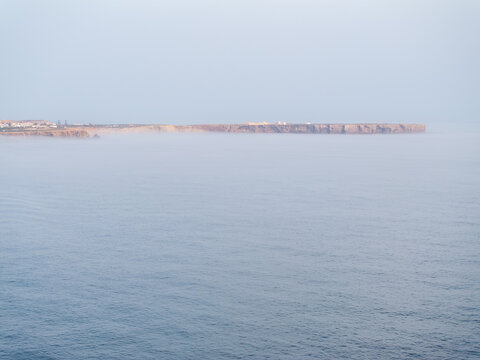 Aerial view of coastal cliffs partially obscured by sea mist rising from the calm ocean in Faro District, Portugal.