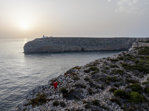 Aerial view of a person in a red shirt standing on a rocky cliff edge overlooking the ocean with a lighthouse on a distant headland at sunset in Faro District, Portugal.