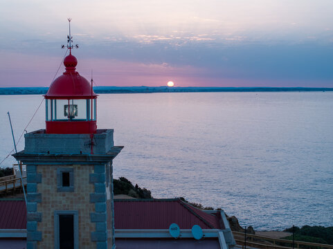 Aerial view of the Ponta da Piedade lighthouse with its red lantern room overlooking the calm ocean during a serene sunset in Faro District, Portugal.