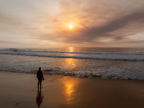 Aerial view of a lone person standing on the sandy beach at sunset with gentle waves rolling onto the shore under a warm glowing sky in Faro District, Portugal.