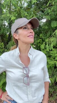 A smiling woman in a baseball cap and white shirt looks up, then gently turns her gaze forward. Her happy, positive expression and casual outdoor style suggest contentment and thoughtfulness amidst lu
