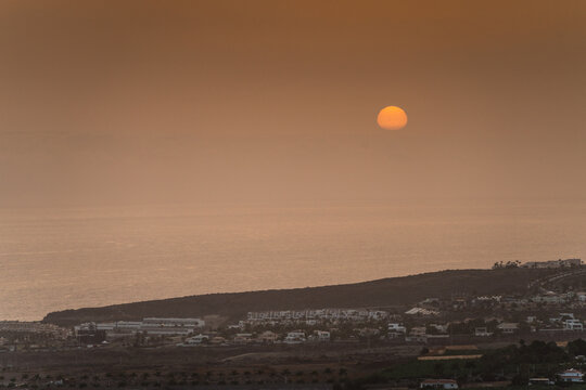 Aerial view of Costa Adeje at sunset with the sun low over the ocean, coastal buildings, and landscape in Costa Adeje, Canary Islands, Spain.