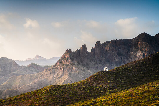 Aerial view of a small white building perched on a rugged hillside surrounded by dramatic mountain peaks under a soft golden hour sky in Costa Adeje, Canary Islands, Spain.