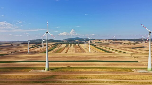 Wind turbines turn in the sun over a large field. The landscape shows rows of crops and flat land. Clear blue skies stretch overhead. It is daytime and the view is open and expansive.