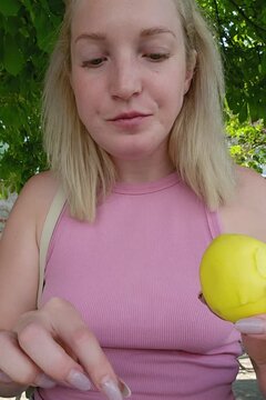 A woman with blonde hair displays a partially bitten lemon-shaped molded shell pastry; her puckered expression highlights the tangy flavor profile, framed by verdant greenery