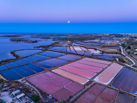 Aerial view of colorful salt pans with pink and blue water reflecting the twilight sky and moon in Faro District, Portugal.