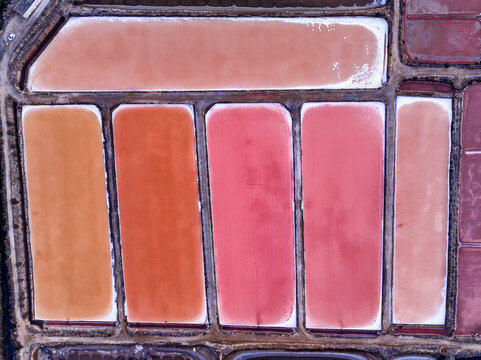 Aerial view of colorful salt pans with varying shades of pink and orange water separated by earthen dikes in the salt marshes of Faro District, Portugal.