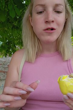 A woman with blonde hair displays a partially bitten lemon-shaped molded shell pastry; her puckered expression highlights the tangy flavor profile, framed by verdant greenery