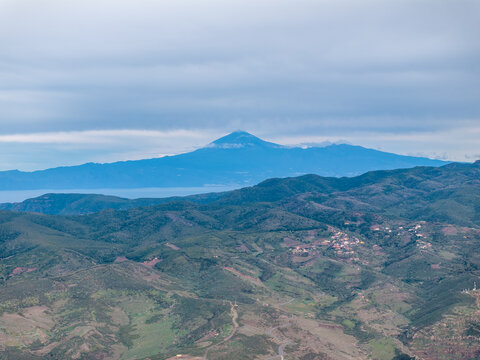 Aerial view of the rugged volcanic landscape of La Palma with rolling hills, sparse vegetation, and distant mountain peaks under a cloudy sky in the Canary Islands, Spain.