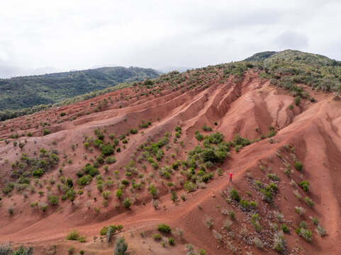 Aerial view of the volcanic landscape of La Palma with red soil, eroded ridges, sparse green vegetation, and a person standing on a trail in the Canary Islands, Spain.