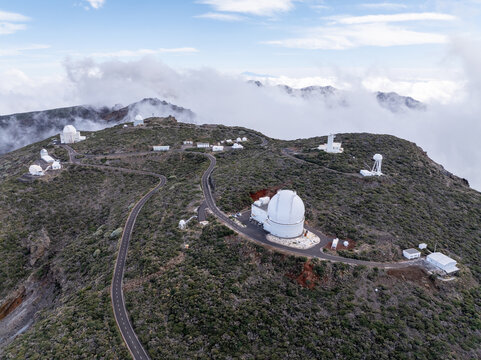 Aerial view of the Roque de los Muchachos Observatory with white telescope domes, winding roads, and volcanic terrain on the mountain ridge in Canary Islands, Spain.