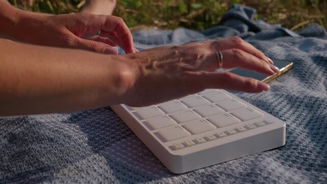 Hands play a white portable MIDI controller on a blanket outdoors in nature during leisure time. Concept of creative freedom, artistic expression, relaxation, and music production in a park.