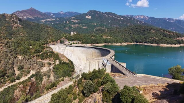 Aerial view of a massive concrete arch dam holding back a blue-green reservoir nestled among forested mountains under a clear summer sky