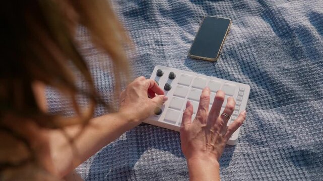 A woman creates music using a portable MIDI drum pad controller on a picnic blanket outdoors. Bright sunlight illuminates the scene near her smartphone during a relaxing outdoor session.
