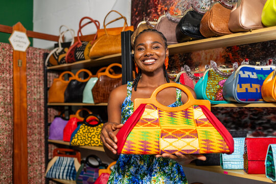 African woman entrepreneur smiles, holding a colorful handcrafted handbag in her small shop