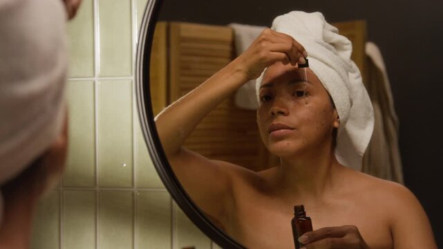 Young Hispanic woman applying facial serum with dropper and massaging skin in bathroom mirror, over shoulder shot. Footage is perfect for skincare ads and beauty marketing campaigns