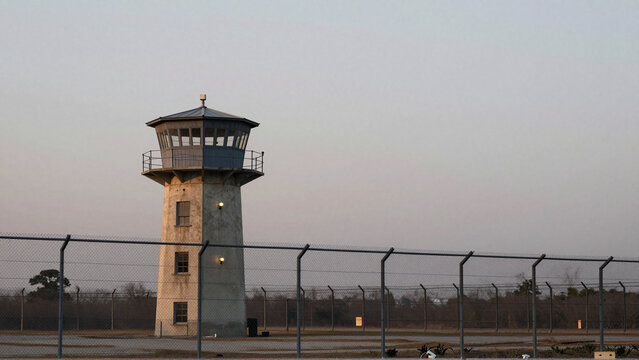 Prison guard tower rises behind layered security fencing under an overcast dusk sky