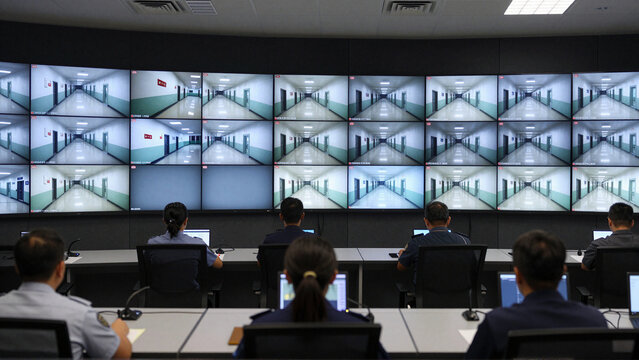 Prison security officers monitor corridor surveillance screens in a central control room