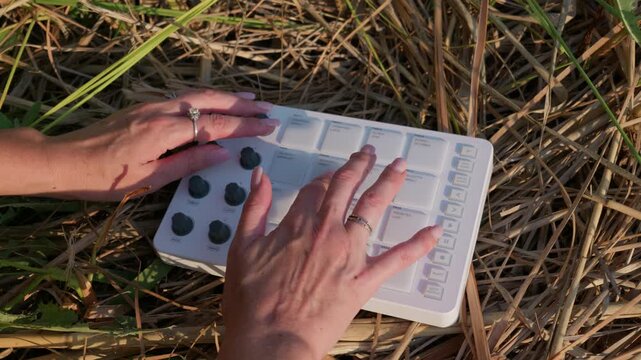 A woman plays a white portable MIDI controller placed on dry straw in an outdoor field. Concept of outdoor music production, creative freedom, and digital technology in nature.