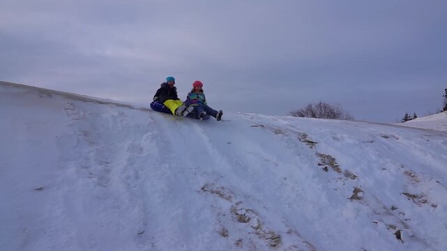 Slow motion two beautiful teenager girls ride from a snow-covered slide on round inflatable wheels.