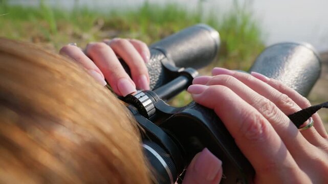 A woman hands with pink nails adjust the focus wheel on a pair of black binoculars outdoors. The close-up shot highlights the optical precision and mechanical details of the gear.