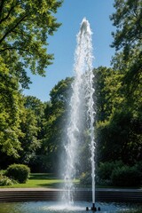 Naklejka premium Close up of tall water fountain in public park. Clear stream shoots up into blue sky. Rich green trees, garden landscape surround stone pool. Refreshing liquid spray creates splash in summer sun for