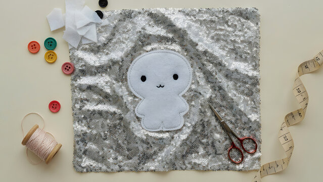 Halloween crafting concept featuring a silver sequin fabric swatch with a cute white ghost cutout and black buttons, surrounded by an orange pumpkin, scissors and colorful sewing materials. Festive, 
