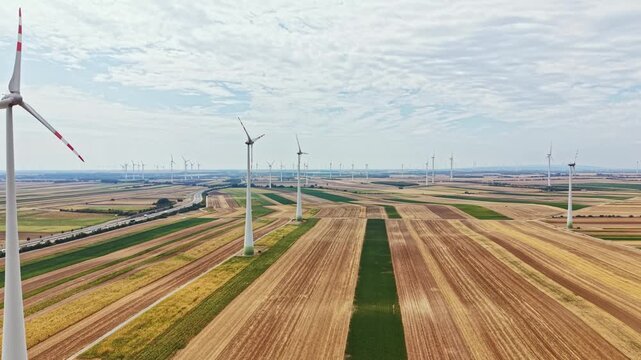Wind turbines rotate in a field during daytime. Green grass and brown soil create patterns on the land. The scene shows renewable energy in use in an open area.