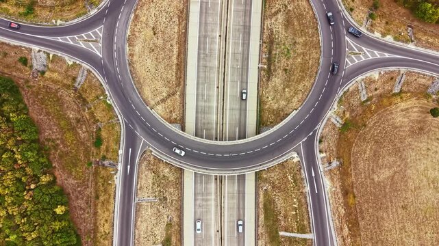 Vehicles navigate a roundabout close to a highway. The surroundings include open fields and green areas. The scene shows clear road markings and smooth traffic movement.