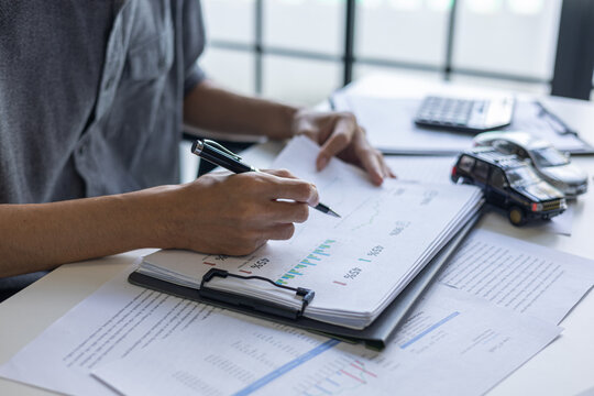 The man is looking at documents to compare car models before making a purchase decision.