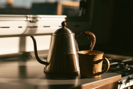 Stainless steel kettle beside ceramic mug in sunlit kitchenette with warm shadows and open counter space for minimalist living