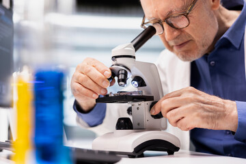 Senior male chemist uses an optical instrument and glass tray to investigate samples in a modern...