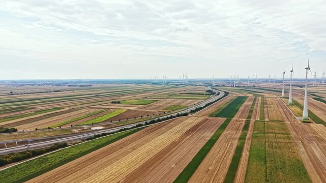 Wind turbines are spread across a vast field where crops grow in neat rows. A road weaves through the landscape. The sky is covered with clouds during the daytime.