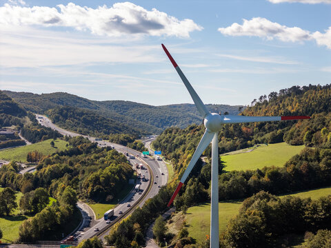 Wind turbine stands tall beside a busy highway with vehicles. Green hills fill the background on a sunny day.