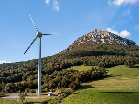 Wind turbines turn in a rural area with green fields and mountains in the background. Clean energy generation is happening here.