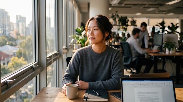 A woman sits at her desk looking out a window into warm sunlight. She takes a mindful break to prevent burnout and set boundaries. Ideal for corporate wellness and mental health content.
