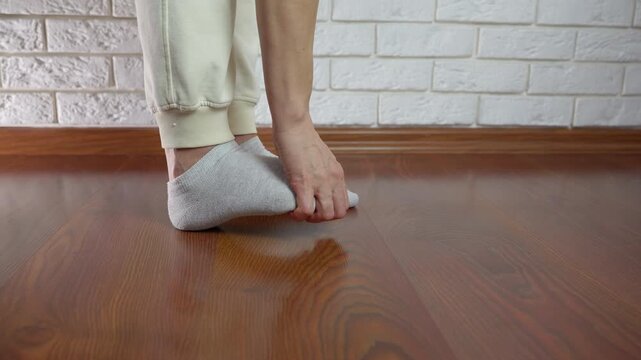 Uncomfortable woman in socks standing on a wooden floor scratching her itchy foot because of a fungal infection, athlete's foot, or an allergic reaction, showing signs of skin irritation